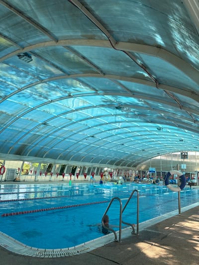 The image depicts an indoor swimming pool facility in Beit Yitzhak-Sha'ar Hefer, Israel. The pool is rectangular with multiple lanes marked by floating lane lines. The water is clear blue. A person is partially submerged in the water near the ladder. The pool is covered by a translucent, curved roof supported by a metal framework, allowing sunlight to filter through and cast dappled shadows. The sides of the pool area are lined with concrete decking, and there are metal railings for pool access. Red triangular flags are visible along one side of the pool. In the background, several people are seated on chairs, and there are trees and other foliage visible through the open sides of the structure. A life preserver with blue and white rings is mounted on a pole near the pool's edge. The overall environment suggests a recreational or training facility.