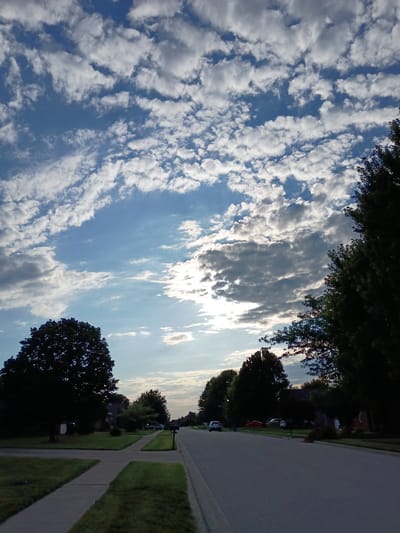 The image depicts a street scene in a suburban neighborhood, likely in Berea, United States of America. The perspective is from the sidewalk, looking down a paved residential street lined with manicured lawns and mature trees. Several cars are visible parked in driveways and along the street. The sky is a dominant feature, filled with a dramatic display of white and gray clouds against a blue backdrop, with hints of sunlight breaking through. The lighting suggests it is either early morning or late afternoon. The overall impression is a peaceful, ordinary day in a residential area.