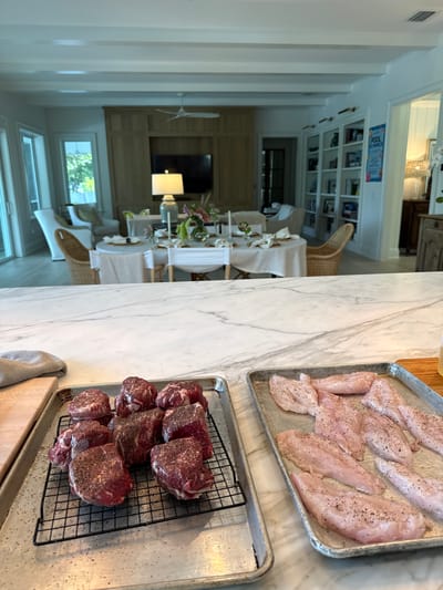 The image shows a kitchen scene with two metal baking trays in the foreground. The tray on the left contains several pieces of raw beef, seasoned with salt and pepper, arranged on a wire rack. The tray on the right holds multiple pieces of raw fish, also seasoned. In the background, a dining room is visible, set with a table covered in a white tablecloth, complete with place settings and floral arrangements. The room features light-colored walls, wooden cabinetry, bookshelves filled with books, and a ceiling fan. Large windows and sliding glass doors offer a view of the outdoors. The overall setting suggests a home in East Naples, Florida, United States of America, preparing for a meal.
