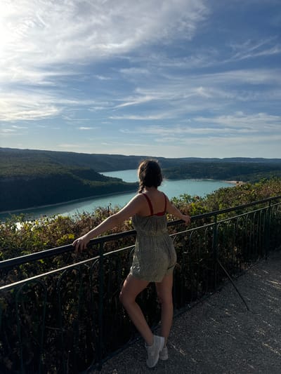 A woman with braided dark hair stands on a gravel walkway, leaning on a black metal railing. She wears a light-colored floral romper and grey sneakers, facing away from the camera towards a vast, turquoise body of water bordered by lush green hills. The sky above is a clear blue with wispy white clouds. The scene suggests a scenic overlook, likely in the vicinity of Moirans-en-Montagne, France, given the characteristic landscape of the Jura region.