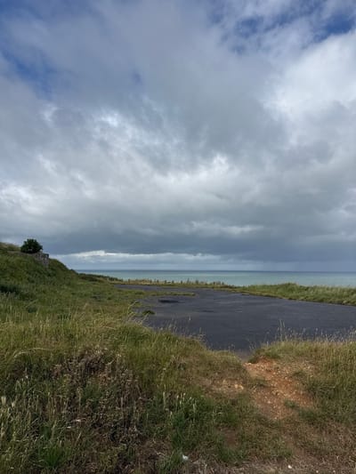 The image depicts a coastal landscape in Octeville-sur-Mer, France, under a heavily overcast sky. The upper two-thirds of the frame are dominated by dense, multi-layered grey clouds, with some darker formations towards the horizon and a few lighter patches hinting at blue sky in the upper left. The horizon line is clearly defined, separating the sky from the sea. The sea appears a muted turquoise-green, exhibiting a choppy surface.

The land mass occupies the lower third of the image, sloping upward from the left and featuring an asphalt-like surface in the foreground and midground. This dark, paved area shows signs of wear, including lighter discoloration and potential cracks. Dense, unkempt vegetation, consisting primarily of tall green and brown grasses and herbaceous plants, frames the asphalt surface and covers the sloping terrain. On the left side, a concrete structure, possibly part of an old bunker or fortification, is partially visible, overgrown with grass. A small, bushy tree with white blossoms grows atop this structure. Along the distant edge of the land, near the sea, a series of vertical posts or stakes is discernible. In the far distance, on the sea, a faint, slender vertical structure (possibly an offshore platform or mast) is visible. The immediate foreground on the right features a small mound of reddish-brown earth partially covered by grass. No individuals are present in the scene.