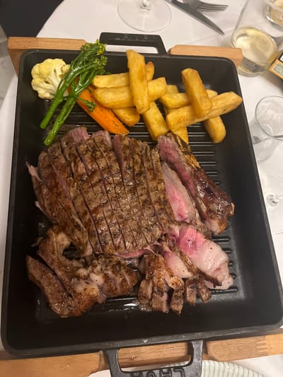 A rectangular, black cast iron grill pan resting on a wooden surface holds a sliced T-bone steak, accompanied by a side of thick-cut fries, broccoli florets, a carrot, and cauliflower. The steak appears to be cooked to different levels of doneness, with some slices well-done and others rare. The surrounding area is a dining table, with cutlery, glasses, and bottles visible. The image was likely taken at a restaurant or private residence in Urbanització La Cometa, Spain, judging by location metadata.