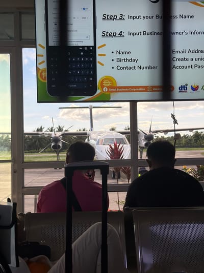 The image shows an airport waiting area, possibly in Del Carmen, Philippines, based on the DTI logo displayed on a screen. Visible through the window is a small aircraft on the tarmac. Inside, people are sitting on waiting chairs, partially obscuring the view of the airplane. A screen displays a digital form, with prompts for inputting business information such as business name, owner's information, email address, name, birthday, and contact number. A digital keyboard is visible on the screen.