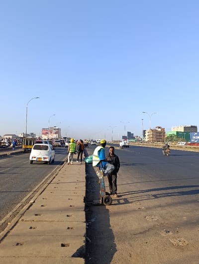 A sunny daytime street scene shows two men standing next to a trolley. One man in a dark jacket stands beside the trolley, while another wearing a yellow hard hat and safety vest is seated on it. Behind them are multiple lanes of a busy highway with cars, trucks, and motorcycles. People walk along the roadside, and buildings are visible in the distance under a clear blue sky.