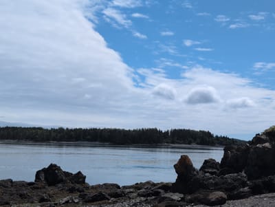 This is a landscape photograph of a coastal scene. In the foreground, there is a rocky shoreline with dark, jagged rocks and some smaller pebbles. The water is calm and a light blue, with some reflections of the sky. In the middle ground, a dense forest of green trees lines the distant shore, creating a dark, solid line against the water. The sky above is a vibrant blue, with large, fluffy white clouds dominating the upper portion of the image, partially obscuring the blue sky in some areas. The image is taken from a low angle, emphasizing the rocks in the foreground and the expanse of water and sky. The location is West Isles Parish, Canada.