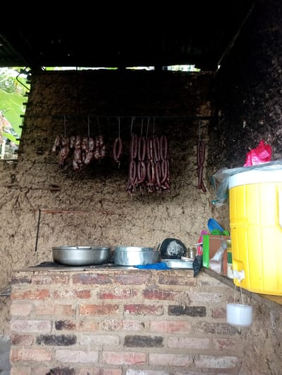 A photograph depicts an outdoor kitchen scene, possibly in a rural setting, featuring a brick cooking area covered with stainless steel bowls and a row of dark sausages hanging from a metal bar above. The sausages are various lengths. A bright yellow cooler sits to the right with a water dispenser. The brickwork is rough and textured, and the walls appear to be made of mud or a similar earthen material. A variety of objects, including a black pot, are arranged alongside the cooler, suggesting a functional kitchen space. The location is inferred to be in Honda, Venezuela, based on the user's prompt.