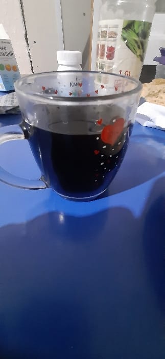 A clear glass mug, decorated with red and white hearts, is filled with a dark liquid. It sits on a glossy blue surface, casting a faint shadow. In the background, out of focus, are a plastic bottle and a cardboard container. The composition is somewhat off-center and tilted, giving a casual, snapshot-like feel.