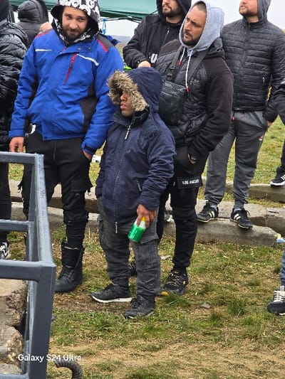 The image depicts a group of six adult males standing outdoors on a grassy area in Borovan, Bulgaria. The sky appears overcast.
In the foreground, a dark grey metal barrier or railing is visible on the left. The ground is covered in sparse green grass and some exposed soil. In the mid-ground, a man of short stature, with dark skin and a dark beard, wears a navy blue hooded jacket with a fur trim on the hood, dark grey cargo pants, and dark sneakers. He holds a green and white bottle in his right hand. To his left, a taller man with a dark beard wears a white patterned hoodie under a blue and red jacket, dark pants, and black boots. He stands with his hands in his pockets and looks towards the camera.
To the right of the central short man, another man with a dark beard wears a light grey hoodie under a black quilted vest and a black long-sleeved shirt. He carries a black shoulder bag across his chest, wears dark pants with "LOS ANGELES" text near the ankle, and dark sneakers with yellow accents. He is looking upwards and to his left. Behind him, another man with a beard wears a black quilted jacket, grey sweatpants, and black and white athletic shoes, with his left hand in his pocket. Partially visible on the far right is another individual wearing dark pants and sneakers.
In the background, blurred trees or distant terrain are visible. A section of a light green tarp or awning is present in the upper left background. Concrete steps or debris are situated behind the individuals on the right.