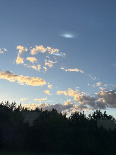 The image shows a sky with wispy clouds illuminated by the setting sun. To the right, there is a dark silhouette of trees. The sky is blue and clear except for the scattered clouds. A light aberration is visible in the upper left corner of the image. The location appears to be Мажэйкаўскі сельскі Савет, Belarus, based on the type of landscape. The image shows a natural environment with vegetation and a visible sky. No people or interactions are seen in the image.