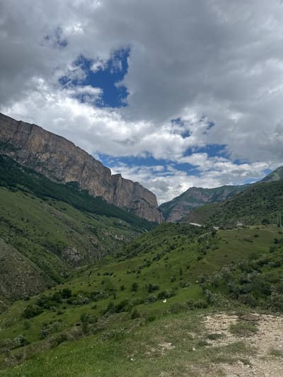 The image shows a vast, rugged mountain landscape under a cloudy sky with patches of blue. In the foreground, a dirt path is visible on the right, covered in sparse grass and small stones. The middle ground is dominated by rolling green hills, dotted with scattered bushes and trees. A winding road can be seen cutting through the hills in the distance. The background features immense, steep rock formations with sheer cliffs. The overall scene depicts a remote, natural mountain environment, likely in or near the city of Verkhnyaya Balkariya, Russia, given the context. There are no people or animals explicitly visible in the image. The weather appears to be overcast with intermittent sunlight breaking through.