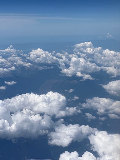 The image is an aerial view of a landscape with a vast expanse of blue sky and fluffy white clouds. Below the clouds, a dark blue, mountainous terrain is visible, with patches of snow on some peaks. There are no visible people, subjects, or objects that would indicate a specific location in the city of Vrellë, Kosovo. The scene depicts natural geographical features from an elevated perspective.