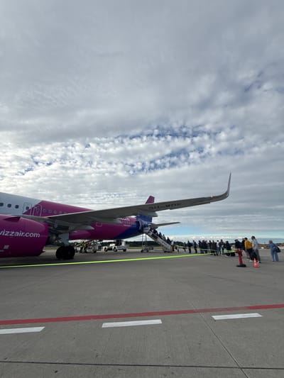 An aerial view of a Wizz Air Airbus A320neo aircraft, registration 9H-WDK, is parked on the tarmac at an airport, likely in or near Künersberg, Germany, as indicated by the context of the prompt. A boarding stairs is extended to the aircraft's front left door, and a line of passengers is queuing to board, some holding luggage. Two ground crew members are visible near the aircraft's front wheel and engine. The aircraft's fuselage is predominantly pink and white, with "wizzair.com" branding visible. The sky is partly cloudy, with a mix of white and grey clouds. The ground is concrete, marked with yellow and red lines. In the background, a treeline is visible, suggesting a rural or suburban setting.