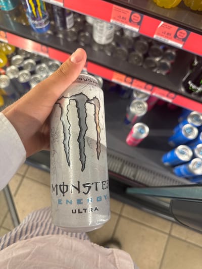 A hand holds a Monster Ultra energy drink can, the silver can displaying the Monster logo. In the background, a cooler shelf is stocked with various energy drinks and other beverages. Red labels indicating prices are visible on the shelf. A section of a person's striped shirt is seen on the right side of the frame.