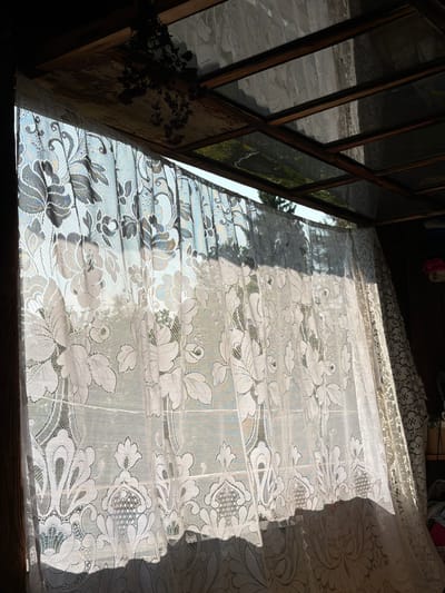 The image depicts an interior view looking out through a lace curtain. The curtain is white and features a floral pattern. Sunlight streams through the curtain, casting shadows and highlighting the intricate lacework. Beyond the curtain, trees and sky are visible, suggesting an outdoor environment. The ceiling above appears to be made of wood with glass panels. The overall impression is one of natural light filtering into a space, possibly a room or sunroom. There are no people or active subjects visible in the scene. The location is not specified as Sylva, Russia.