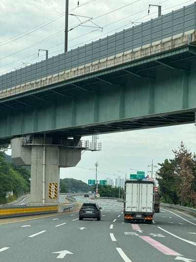 The image captures a view from a vehicle driving on a highway in Yangsan-si, South Korea. The foreground shows a multi-lane asphalt road with white directional arrows painted on the surface. Two vehicles are visible: a dark-colored SUV in the right lane, and the back of a white box truck in the left lane.
In the background, a massive elevated structure, likely a highway overpass or railway bridge, dominates the scene. The underside of this structure is painted a dark green, supported by large concrete pillars. Above the overpass, overhead power lines for an electric train are visible against a cloudy sky.
Road signs are present, including a green directional sign indicating "Gyeongju" and "Ulsan" in Korean and English, along with other Korean signage on a yellow and black warning sign attached to a concrete pillar. Trees and vegetation line the sides of the highway, with some buildings visible in the distance on the right. The overall impression is a typical highway scene in South Korea, characterized by extensive infrastructure and ongoing traffic.