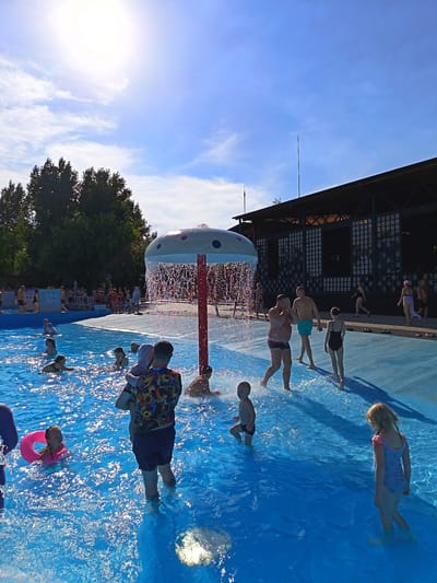 The image depicts a sunny day at an outdoor swimming pool facility in Yarovoye, Russia. In the foreground, the bright blue water of the pool is filled with many people. A young child is being held by an adult in the water. A pink inflatable ring floats near a young girl in the pool. Further into the pool, several other individuals, including children and adults, are visible swimming and playing.
A mushroom-shaped water feature with water spraying down from it is positioned in the middle of the pool. To the right of the water feature, a group of people are standing or walking on a blue tiled area surrounding the pool. Among them is a young girl in a swimsuit and a young boy in swim trunks. In the background, there is a large, dark-colored building with a distinctive architectural style, likely a part of the pool complex. Trees and a bright blue sky with scattered white clouds complete the scene.