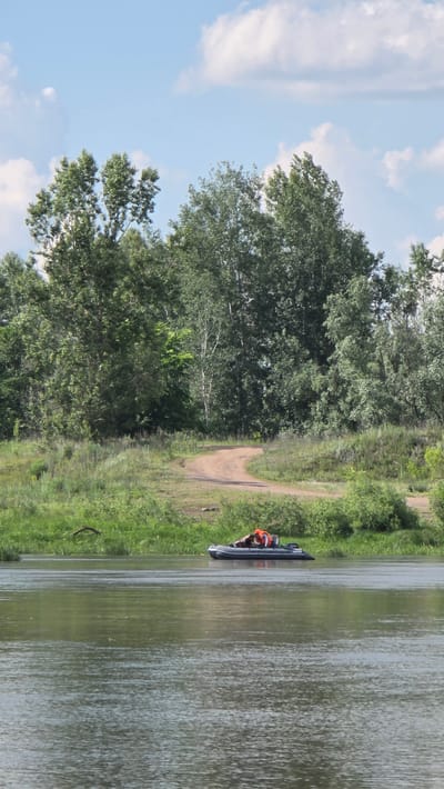 An inflatable boat with a person wearing an orange life vest is on a calm river in Дарьино, Russia. The boat is dark gray and appears to be powered by a small outboard motor. The person is seated and looking forward. The riverbank behind the boat is lined with green vegetation and trees. A dirt road winds up a gentle slope behind the vegetation. The sky is blue with scattered white clouds.