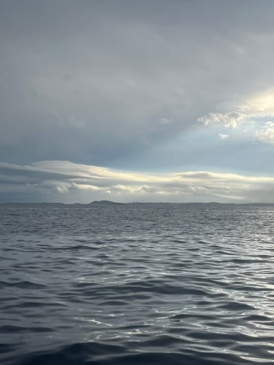 A calming seascape showcases a contrast between sky and water. Turbulent, choppy waves fill the right side, reflecting sunlight with a shimmering effect. To the left, a dramatic sky features a mix of dark, heavy clouds and patches of brighter, diffused sunlight. A narrow strip of land faintly separates the sky and the water, creating a distinct horizon line. The overall scene evokes a sense of vastness and the power of nature.