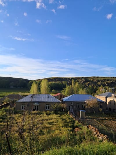 The image shows a rural landscape in Artsvakar, Armenia, under a bright blue sky with scattered white clouds. In the foreground, a cluster of houses with various roof types, including corrugated metal and tiled roofs, are nestled amongst green, undulating terrain. Some houses appear to be single-story structures with visible windows and doorways. The vegetation is lush, with grassy fields and scattered trees, some of which are bare and leafless, suggesting either early spring or late autumn. In the middle ground, a gentle slope is covered with trees, and further back, a higher, rocky hill forms the horizon. The overall impression is of a peaceful, rural setting.