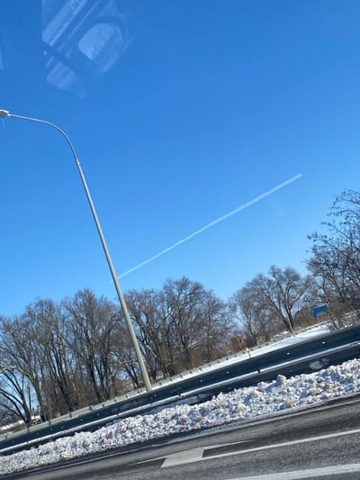 The image displays an outdoor winter scene in the city of Stepnoe, Russia, captured from an elevated perspective, likely from inside a moving vehicle. The upper half of the frame is dominated by a clear, bright blue sky containing a prominent, thin white contrail extending diagonally from the upper right towards the lower left. A tall, grey metallic lamppost with a curved arm rises from the lower left quadrant towards the center-top. A faint, indistinct reflection, consistent with an interior vehicle structure, is visible in the upper left corner of the sky.
The midground features a dense line of leafless deciduous trees with dark branches, forming a horizontal band across the scene. Below the trees, a metallic guardrail runs parallel to the bottom edge of the image, separating the tree line from the foreground. Accumulations of dirty snow are visible on the ground immediately behind the guardrail and alongside the road. The foreground consists of a dark asphalt road surface with a discernible white marking. In the distant background, to the right, a blue and yellow sign is partially visible, and a faint light-colored building structure can be seen to its left, behind the trees. No individuals are present in the scene.