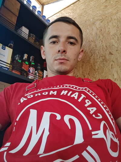 The image shows a young adult male, appearing to be in his mid-20s, seated in what seems to be a storage or retail area. He is wearing a red t-shirt with a large white logo featuring the letter "M" and text that reads "CAPTAIN MORGAN". Behind him, shelves are visible, stocked with various bottles, some of which appear to be alcoholic beverages, and blue cups. The background wall is made of oriented strand board (OSB). The lighting suggests an indoor setting, possibly a bar or a shop. The overall impression is a casual, candid photograph. The location is identified as Shkurupiivka, Ukraine.
