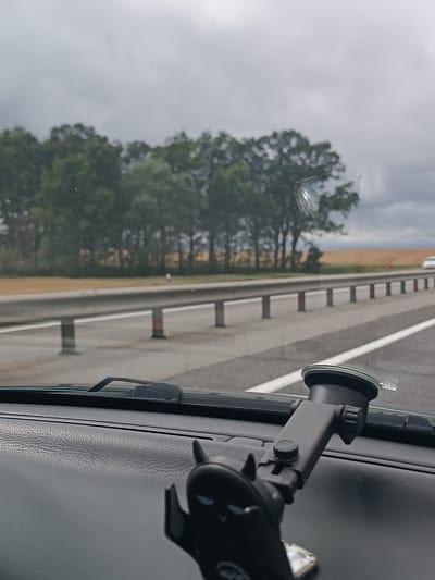 The image captures a view from inside a vehicle, looking through the windshield. The dashboard is visible in the foreground, with a phone mount attached to it. The mount is dark grey and has a suction cup base. Beyond the windshield, a highway is seen with a metal guardrail running along the side. Trees line the roadside, and a field of dry grass or crops is visible in the distance. The sky is overcast with grey clouds. The scene appears to be a highway in the vicinity of Верцялішкаўскі сельскі Савет, Belarus, based on the metadata.