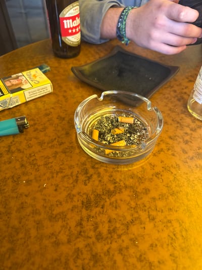 The scene depicts a tabletop in La Lastrilla, Spain, observed from an overhead perspective, likely within a bar or cafe setting.
**Environment:** The table surface is a mottled orange-brown, showing some wear and minor specular reflections. The ambient lighting appears warm, illuminating the objects on the table.
**Objects and Subjects:**
1. **Ashtray:** A clear glass, circular ashtray is centrally positioned, containing a substantial accumulation of dark ash and four orange-filtered cigarette butts.
2. **Cigarettes:** A yellow pack of "Camel" brand cigarettes lies to the left of the ashtray. The visible side features an image of a woman and a Spanish health warning: "El humo del tabaco contiene más de 70 sustancias cancerígenas."
3. **Lighter:** A turquoise blue disposable lighter is situated to the left of the cigarette pack.
4. **Beer Bottle:** A dark brown glass bottle of "Mahou Cinco Estrellas" beer, identifiable by its red, white, and gold label, stands in the upper left section of the table. Its contents are not visible.
5. **Empty Plate/Tray:** A small, square, dark brown or black plate with a slightly raised rim rests behind the ashtray. It appears empty.
6. **Person:** A person's forearm and hand are visible in the upper right. The person wears a long-sleeved gray shirt and a green and black braided bracelet on the wrist. The hand is gripping a dark, indistinct object, possibly a mobile phone.
7. **Additional Bottle:** A clear glass bottle, partially visible on the far right, appears empty or contains a transparent liquid, with an unidentifiable label fragment.
**Actions/Interactions:** The arrangement of smoking paraphernalia (ashtray with butts, cigarette pack, lighter) and a beer bottle suggests recent or ongoing consumption of both tobacco and alcohol. The visible hand is engaged with a personal item.