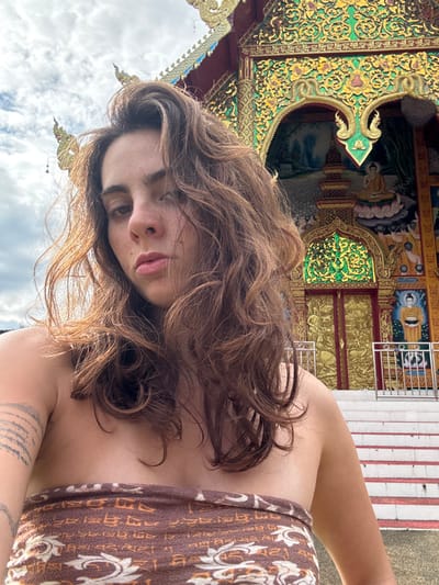 A female subject poses for a self-portrait in front of a richly adorned Buddhist temple in Ban Mae Na Chalong, Thailand. The woman has long, wavy brown hair, which appears sun-kissed, and wears a strapless brown garment featuring a patterned design. A tattoo consisting of multiple horizontal lines is visible on her left upper arm. Her expression is neutral as she looks directly into the camera. The temple facade behind her is highly ornate, showcasing intricate gold leaf patterns, red accents, and green embellishments. Visible architectural elements include a multi-tiered roof with golden finials (chofas) and a prominent central entryway. This entryway features golden double doors, embossed with figures, situated above white stairs with silver railings. Above the doors, within an arch, a painting depicts a seated Buddha figure on a lotus, and to the right, a standing Buddha is depicted within another panel. The sky above is overcast with visible white and gray clouds.