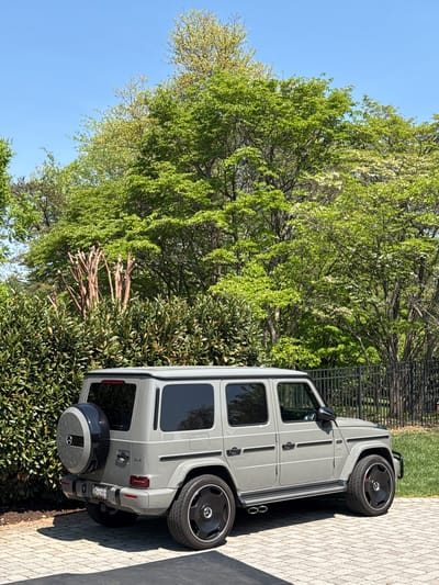 A light gray Mercedes-Benz G-Wagen (G-Class SUV) is prominently featured, parked on a paved driveway made of light-colored paver stones. The vehicle is seen from its passenger side, with the front facing away from the viewer towards the left. It has distinct black multi-spoke wheels with Mercedes-Benz star logos in the center, and its side-exit exhaust pipes are visible beneath the rear passenger door. A "V8 BITURBO" badge is present on the front fender. The rear of the vehicle shows a covered spare tire mounted on the door, also featuring the Mercedes-Benz star logo. A partially visible license plate on the rear appears to be from the United States.
The car is situated outdoors on a clear, sunny day, suggested by the bright light and distinct shadows. The background is dominated by lush green trees with fresh, light-green foliage, indicating springtime or early summer. A vibrant blue sky is visible above the trees. Behind the car, a black metal fence separates the driveway from a grassy area. In front of the rear of the car and extending into the lower frame, there are dense green bushes. The overall setting suggests a well-maintained residential or suburban environment, consistent with the provided location context of Potomac, United States of America. No people are visible in the image, and no specific activity is taking place beyond the car being parked.