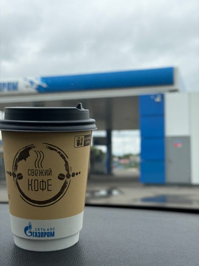 A close-up, eye-level shot reveals a disposable coffee cup positioned on a dark, textured vehicle dashboard, with a blurred Gazprom gas station in the background under a cloudy sky in Совхозный, Russia. The cup, predominantly light brown/kraft paper, features a black plastic lid. Its design includes a distressed circular emblem with the Russian text "СВЕЖИЙ КОФЕ" (Fresh Coffee), flanked by stylized coffee beans and steam rising from a cup graphic. Above this, a small icon with a thermometer and a hot beverage symbol is accompanied by the Russian warning "ОСТОРОЖНО ГОРЯЧО!" (Caution Hot!). The bottom of the cup displays the blue Gazprom logo and the text "СЕТЬ АЗС ГАЗПРОМ" (Gazprom Gas Station Network) on a white band. The background shows the blue and white structures of a gas station canopy with a partial "GAZPROM" logo visible on the left, and a prominent blue pillar on the right. No individuals are present in the image.