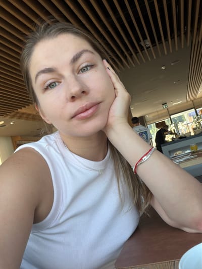 A young woman with light brown hair, green eyes, and fair skin takes a selfie. She is wearing a white ribbed tank top and a silver necklace with a pendant. Her right hand is resting on her cheek, and she has a red and silver bracelet on her left wrist. In the background, a ceiling with wooden slats is visible, along with blurred figures and a buffet area. The lighting suggests an indoor setting during the daytime. There is no information about the location in Žunovnica, Bosnia and Herzegovina.