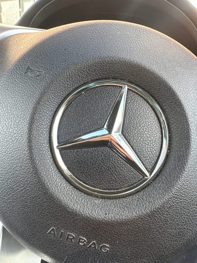 The image displays a close-up, high-angle view of the central portion of a black Mercedes-Benz steering wheel. Dominating the center is the Mercedes-Benz three-pointed star logo, rendered in chrome or polished metal, enclosed within a circular chrome ring. The steering wheel's surface is a textured black material, likely plastic or leather, characteristic of an airbag module. Below the central logo, the word "AIRBAG" is embossed in capital letters. To the upper left of the logo, a small, stylized horn symbol (bugle) is embossed. Reflections of an unseen light source are visible on the metallic logo. No individuals are present in the frame. The immediate environment is the interior of a vehicle. The specific location in Easingwold, United Kingdom, cannot be determined from the visual information provided.