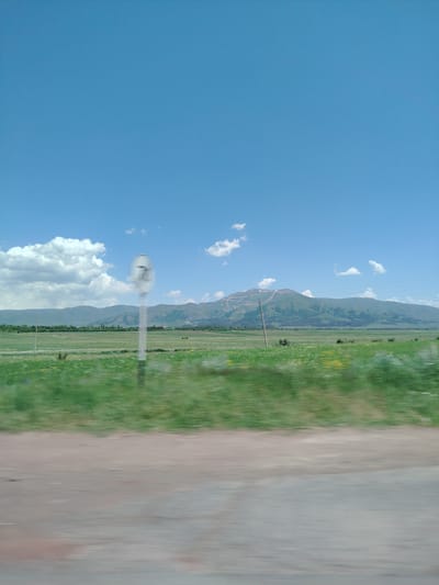 The image captures a roadside view of a landscape, likely taken from a moving vehicle, given the motion blur. In the foreground, a dirt or gravel road is visible with significant motion blur. Beyond the road, a vibrant green field stretches out, dotted with small yellow flowers and appearing somewhat blurred as well. In the middle ground, a white pole with a circular sign bearing the number "7" is prominently displayed, slightly blurred. Further in the distance, a range of rolling green mountains forms the horizon, with some patches of snow or light-colored rock visible on their slopes. The sky is a clear, bright blue with a few scattered white clouds. The overall impression is a rural or semi-rural setting under a sunny sky. There are no people or animals clearly visible in the image. The location is identified as Kuchak, Armenia.