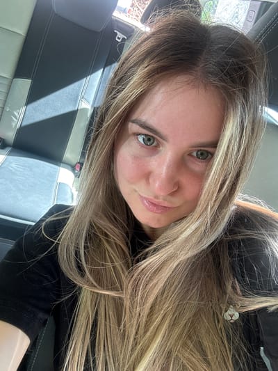 A woman with long, blonde hair is taking a selfie in a car. She is wearing a black shirt and has green eyes. Sunlight is casting shadows on the car seats behind her. There is a small earring with a cat's face visible on her right side.