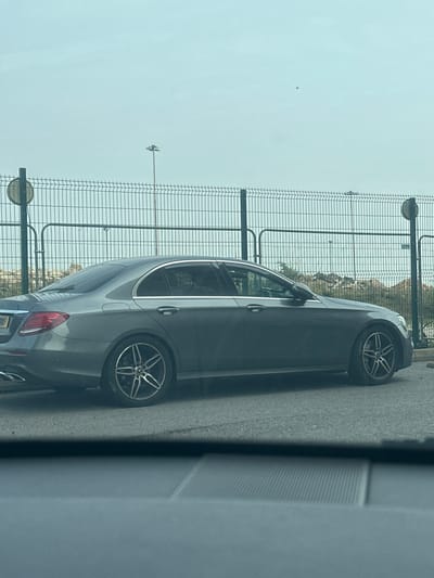 A dark grey Mercedes-Benz E-Class sedan (W213 generation) is parked facing right on an asphalt road surface. The vehicle features multi-spoke alloy wheels and a partially visible UK license plate on its rear. A single individual is discernible as a silhouette in the driver's seat. In the background, a tall, dark green metal mesh security fence with horizontal support bars spans the frame, featuring multiple vertical posts and two visible gated sections. A circular sign is affixed to a fence-line pole on the left. Beyond the fence, irregular mounds of earth or debris are present, with a tall, multi-head floodlight tower visible in the distant background under a uniformly light grey, overcast sky. The image appears to be taken from inside another stationary vehicle, with a blurred dashboard or interior element occupying the lower foreground. This scene is consistent with an industrial or commercial area within Gillingham, United Kingdom, such as those adjacent to port facilities or within business parks.
