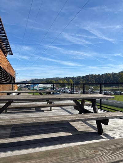 The image captures an outdoor terrace or elevated platform overlooking a semi-industrial landscape in Lièpvre, France, under a clear blue sky. In the foreground, three weathered wooden picnic tables with attached benches are positioned on a wooden deck, casting distinct shadows indicating bright sunlight. A black metal railing with glass panels separates the terrace from the area beyond. To the left, a modern building section with horizontal wooden paneling and dark grilles is visible. The midground features a large asphalt parking lot populated with numerous parked cars of various colors. Beyond the parking lot, several industrial-style buildings, including one with a blue roof, are present, interspersed with green grassy areas and a paved road. The background is dominated by densely forested hills. The sky is a vibrant blue with sparse, wispy white clouds and several visible contrails. Multiple power lines diagonally traverse the upper left sky, originating from a utility pole partially visible on the far left. The scene is devoid of human activity.