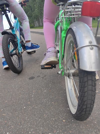 Two individuals are riding bicycles on an asphalt road. The person on the left is riding a light blue bicycle, and the person on the right is riding a bright green bicycle. Both individuals are wearing colorful leggings and athletic shoes, suggesting a casual outdoor activity. The environment appears to be a suburban or rural setting, with trees and foliage visible in the background. The lighting suggests it is daytime. The specific location provided, "Луч Снт (белые Столбы), Russia," indicates a garden non-profit partnership area, which aligns with the observed environment. The image captures a moment of active recreation.