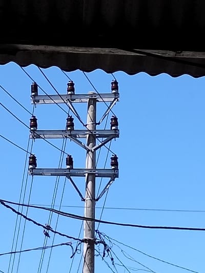 A utility pole stands tall against a clear blue sky. Three horizontal bars extend from the pole, each supporting three brown ceramic insulators. Electrical wires run along the pole, connected to the insulators. A corrugated roof section obscures the view from the top of the image. At the base of the pole, more wires hang loosely.