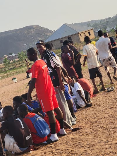 A wide-angle, eye-level shot depicts a group of young men in Ibo, Nigeria, engaged in an outdoor activity. The men are positioned on a reddish-brown dirt surface. They are wearing a variety of clothing, including sports jerseys, shorts, t-shirts, and socks. Some have backpacks near them. A building with a tin roof and a partially constructed brick structure is visible in the background. Further in the distance, a large, rocky mountain formation is seen against a hazy sky. The environment appears to be a somewhat arid, grassy field.