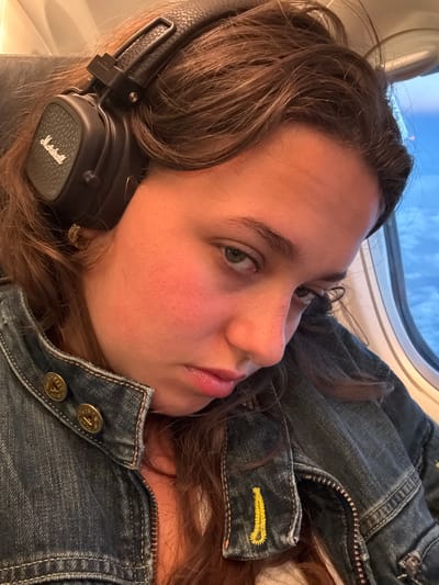 A young adult female with medium-brown hair is depicted in a close-up, seated within a vehicle, likely an aircraft due to the distinct oval window shape. She is wearing black over-ear Marshall headphones on her head, with the brand name visible in white cursive on the left earcup, which features a textured surface. Her hair is slightly disheveled around the headphones. Her complexion is pale, with noticeable redness on her cheeks. She has hazel or light green eyes, which are directed towards the viewer with a somewhat contemplative or melancholic expression, and her lips are slightly parted. A small, gold-toned hoop earring is visible in her right earlobe. She is dressed in a dark wash denim jacket, partially unzipped to reveal a light-colored undershirt. The jacket features two brass-toned buttons on the collar; one clearly displays "G-STAR" and "RAW" text with "0620" below it. A decorative yellow stitched detail, resembling a stylized loop, is present on the right cuff of the jacket sleeve. The background features a clear blue sky with white, fluffy clouds seen through the vehicle's window, indicative of high altitude. A faint red light is visible in the distant sky outside the window. The scene is captured in Arilje Municipality, Serbia.