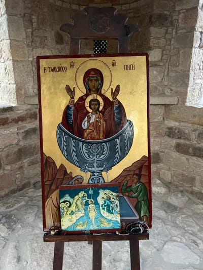 This media file displays an Eastern Orthodox icon of the Theotokos (Mother of God) with Christ, situated within a stone structure, likely a church or chapel in Kolossi, Cyprus. The icon, painted on a gold background with a red border, depicts the Virgin Mary holding the Christ child. They are enclosed within a stylized representation of a chalice or ciborium. The figures are rendered in traditional Byzantine style with halos. To the right of the main icon, a smaller icon depicts a scene with multiple figures, also in a religious context. A man in a green robe sleeps on a brown surface to the left of the smaller icon. Several beaded necklaces, possibly prayer beads or votive offerings, are draped over the wooden easel supporting the icons. The background consists of rough-hewn stone walls and arches, characteristic of ancient religious architecture. Visible Greek text on the main icon includes "Η ΖΩΟΔΟΧΟΣ ΠΗΓΗ" (The Life-Giving Spring) and abbreviations for "MP ΘΥ" (Mother of God). The overall impression is one of reverence and spiritual significance within a historic setting.