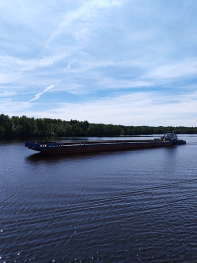 This image captures a wide-angle view of a long, flat-decked cargo barge, colored dark reddish-brown on its hull and dark blue on its upper edges, being propelled by a smaller white and blue tugboat from its stern. The vessels are moving rightward across a body of dark blue water, which exhibits small ripples, surface reflections, and a visible wake trailing the tugboat. The scene is located within the city of Mitino, Russia. A dense line of dark green trees forms the far bank of the waterway, stretching horizontally across the middle ground. Above, the sky is clear and bright blue, interspersed with sparse, wispy white clouds and a distinct, elongated contrail or cirrus cloud diagonally crossing the upper portion. The lighting indicates a bright, sunny day. No individuals are visibly present on the barge or tugboat.