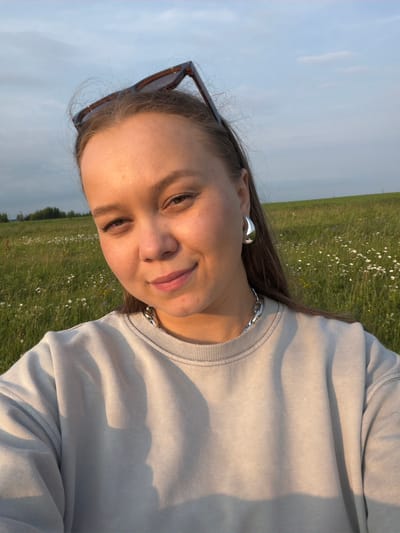 A young woman with blonde hair, wearing a light grey sweatshirt, large silver hoop earrings, and a chunky silver necklace, smiles at the camera. Sunglasses are perched on top of her head. She is in a grassy field dotted with small white flowers, with a line of trees in the distance. The sky is blue with some clouds. The lighting suggests it is late afternoon or early evening.