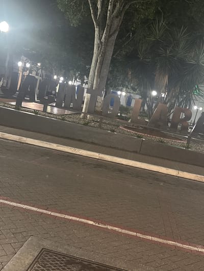 The image depicts a large, three-dimensional metal sign spelling out "JACINTO LARA" at night. The sign is situated in an urban environment, possibly a public square or park. The letters are dark, appearing to be made of metal and are illuminated by surrounding lights, casting a faint glow. Behind the sign, several palm trees and other foliage are visible, along with streetlights and building facades. In the foreground, a paved road with brick-like patterns is visible, marked by a white and red stripe. A storm drain cover is also present in the lower left corner. The overall scene suggests an outdoor public space in a city at night. The sign itself references Jacinto Lara, a historical figure associated with Venezuela, and the city of Barquisimeto, where there is a prominent monument and plaza named after him, making it highly probable that this is the Plaza Jacinto Lara in Barquisimeto, Venezuela.