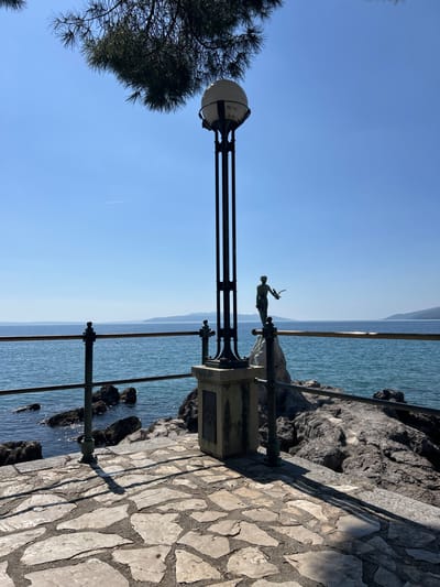 The image captures a sunlit coastal scene in Opatija, Croatia, from the perspective of a paved stone walkway. A prominent black metal lamppost stands on the right side of the path, featuring a spherical light fixture at its top. Attached to the lamppost's base is a bronze statue of a slender female figure, commonly identified as the "Girl with the Seagull," holding what appears to be a bird in her outstretched hand, facing the sea.
To the left of the walkway, a metal railing runs along the edge, providing a barrier above a rugged, rocky coastline. The calm, blue-green waters of the Adriatic Sea stretch out to the horizon. In the distance, faint landmasses or mountains are visible across the water under a clear, bright blue sky. Part of a dark green pine tree is visible on the far left, framing the scene.
The strong, bright sunlight indicates either mid-morning or late afternoon, as evidenced by the long shadows cast by the lamppost and railing across the stone-paved path. The lamppost base features an ornate, possibly floral, emblem, but no legible text is visible. The scene evokes a peaceful and picturesque coastal promenade.