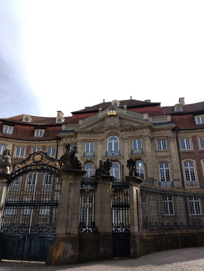 The image shows the Erbdrostenhof in Münster, Germany. The building is a Baroque palace, three stories high, with a red-tiled roof and dormer windows. The facade is made of light-colored stone and brick. A large ornate black metal gate stands in front of the building with stone pillars topped with statues. The sky above is pale blue with some clouds. No people are visible in the image. The setting is a clear day in an urban environment.