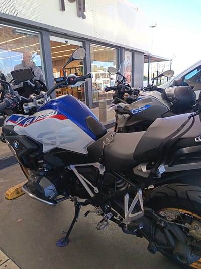 Two BMW GS adventure motorcycles are parked outside a modern building with a dotted facade and glass entrances in Zárate, Argentina, during the daytime under a clear sky.

The prominent motorcycle in the foreground is a BMW R 1250 GS, featuring a distinctive white, blue, and red "HP" livery. "HP R1250" and a large "GS" logo are visible on its tank, along with the "BMW" emblem on its side panel. It has crash bars, a kickstand resting on a purple pad, and its rear wheel shows golden spokes. A digital display mount is attached to its handlebars.

Behind the R 1250 GS, another BMW GS motorcycle is parked, colored grey with blue "GS" graphics on its tank. A black motorcycle helmet rests on its seat. Partially visible behind these motorcycles is a white van.

Through the glass door of the building, a man with short grey hair is seen reflected on the front motorcycle's windscreen and partially visible behind it. He appears to be inside the building, which has bright interior lighting and what looks like shelves or displays. A faint "FUJI" text is visible on a poster inside the building. The ground is concrete, with yellow wheel chocks near the front motorcycle's engine guard. The overall scene suggests a stop during a journey, possibly at a service station or a store.