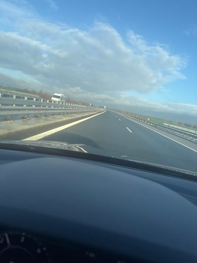 The image depicts a daytime scene from the interior of a vehicle, looking forward through the windshield onto a multi-lane highway in Воденичане, Bulgaria. The vehicle's dark-colored dashboard occupies the lower portion of the frame, showing a glimpse of a speedometer or control panel on the far lower left. The asphalt highway surface, marked with white solid and dashed lines, appears damp or wet, reflecting light in some areas. A metal crash barrier with red reflectors extends along the left side of the highway, separating the road from an embankment featuring sparse, deciduous vegetation. In the distance on the left, a white van, possibly a Mercedes-Benz Sprinter, is visible traveling in the opposite direction. The right side of the highway borders green fields. The sky above is partly cloudy, with significant patches of blue sky visible amidst white and grey cloud formations. The overall environment is open and rural or exurban, under conditions suggesting a recent or ongoing period of damp weather.