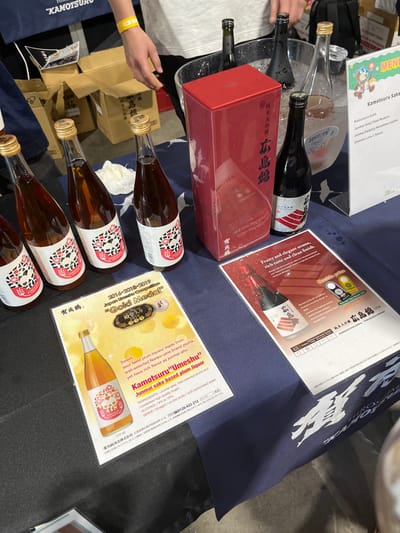 A table covered in a dark blue tablecloth with a white logo in the bottom right corner. On the table are four bottles of Japanese plum wine, labeled "Kamotsuru". Two of the bottles are standing upright, and two are lying on their sides. The bottles are all 720 ml. Next to the bottles, there is a red box with a Japanese label, a small brochure, and a large colorful brochure. The large brochure is advertising a "Gold Medal" winning "Kamotsuru Umeshu" Junmai sake based plum liqueur. The small brochure has a Japanese label and a photo of a bottle of plum wine, "Doubutsu park". The table is located at the Hamilton Quarter Markets in Hamilton, Australia, at the coordinates -27.439, 153.075. The photo is taken from a slightly elevated perspective, looking down at the table. The environment is bright and well-lit.