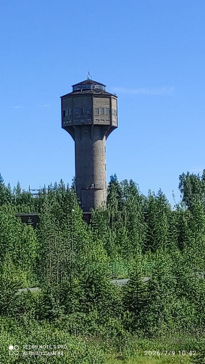 A concrete water tower stands tall against a clear blue sky, surrounded by a dense forest of green trees. The tower has a distinctive octagonal shape at its top, with several windows visible. The base of the tower is obscured by the foliage of the surrounding trees. The lower part of the image shows a path or road winding through the trees, and a small section of a building can be seen to the left of the tower, partially hidden by the trees. A timestamp and camera information are visible in the lower right and left corners of the image, respectively. The location is Otanmäki, Finland.
